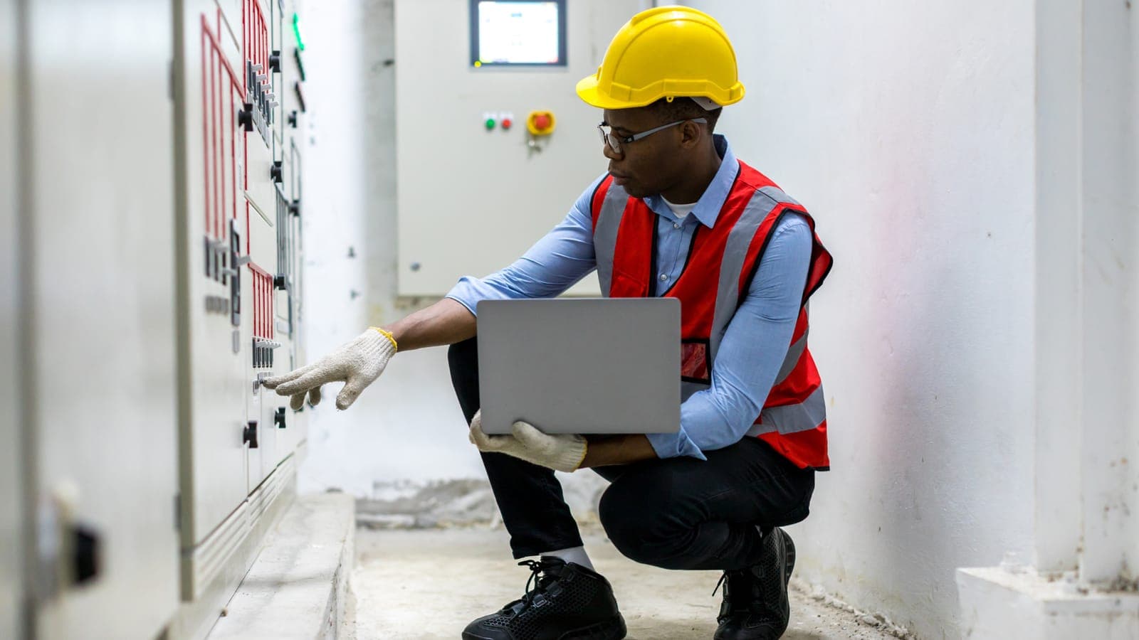 Technician in hard hat and safety vest checking electrical switchgear with a laptop